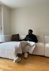 Sötét dzsekis, világos nadrágos férfi fehér szekcionált kanapén ül, leopardmintás párnával egy minimalista szobában.