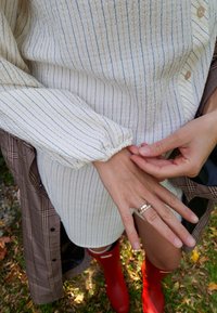 Camisa blanca de manga larga con rayas marinas, acompañada de una capa exterior de cuadros. Botas de goma rojas y tres anillos en la mano.