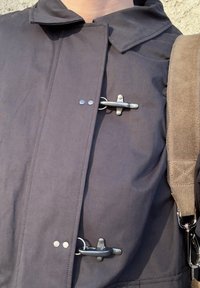 Cappotto grigio scuro resistente all'acqua con chiusure a fibbia, collo strutturato e tessuto liscio con una lucentezza sottile. Tracolla regolabile visibile.