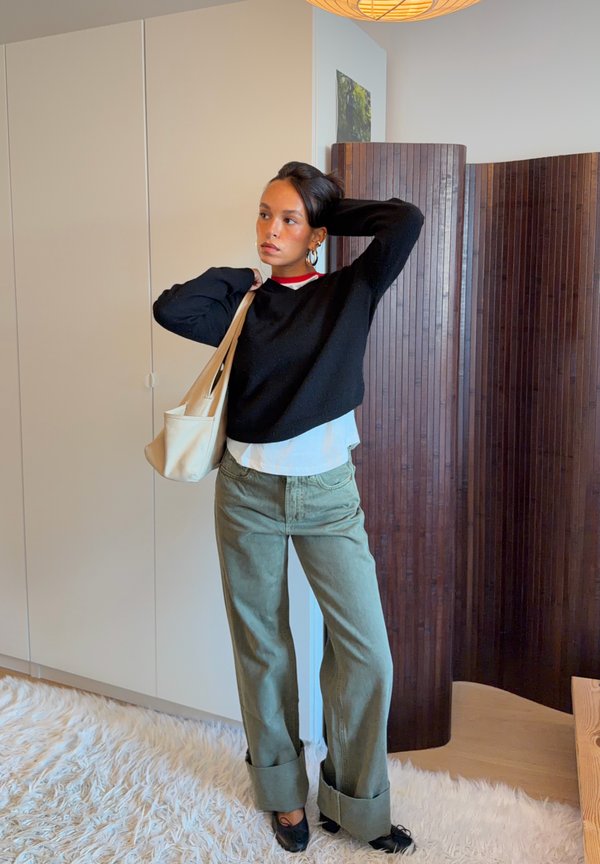 Mujer de pie en el interior con un jersey negro, camisa blanca, pantalones verdes de pierna ancha, zapatos negros y sosteniendo un bolso tote beige sobre su hombro.