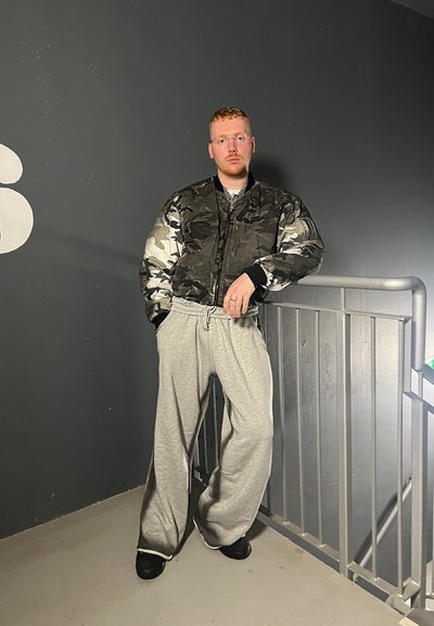 Chaqueta bomber de camuflaje, pantalones de chándal grises y zapatos negros. Texturas suaves con un ajuste relajado. Pared gris y barandilla al fondo.