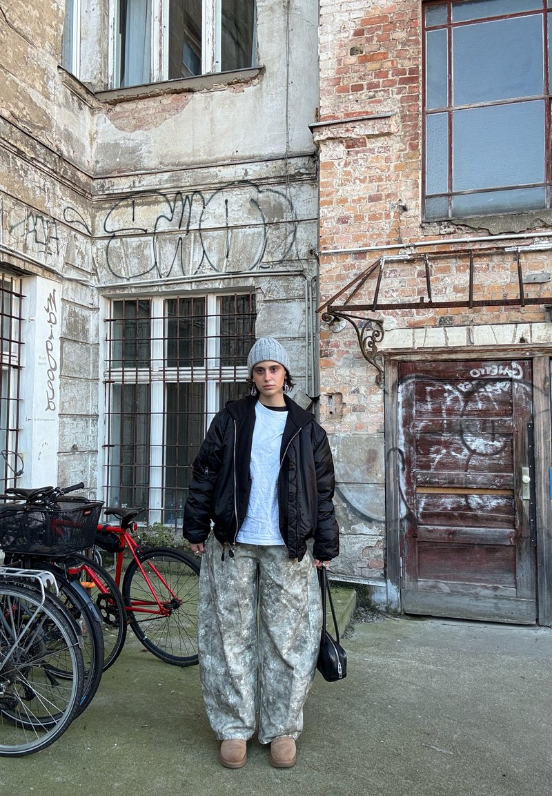 Casaco acolchoado preto, t-shirt branca, calças largas bege com padrão, chinelos bege, gorro cinzento, em pé perto de bicicletas e de uma parede de tijolos.