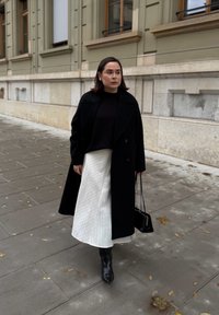 Sort kåbe, mørk sweater og hvidt midi-nederdel med striber. Sorte knælange støvler og lille sort håndtaske. Urbant miljø med belagt underlag.