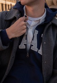 Pilka ribuota striukė, dėvima ant tamsiai mėlynos spalvos džemperio su dideliu „A“ logotipu, po apačia baltas marškinėlių apykaklė, rankose keli žiedai.