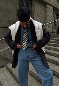 Μαύρο σακάκι, τζιν πουκάμισο, ανοιχτό μπλε τζιν, ριγέ γραβάτα με καφέ λεπτομέρειες, λευκό πουλόβερ ριγμένο στους ώμους, σκούφος, γυαλιά ηλίου.