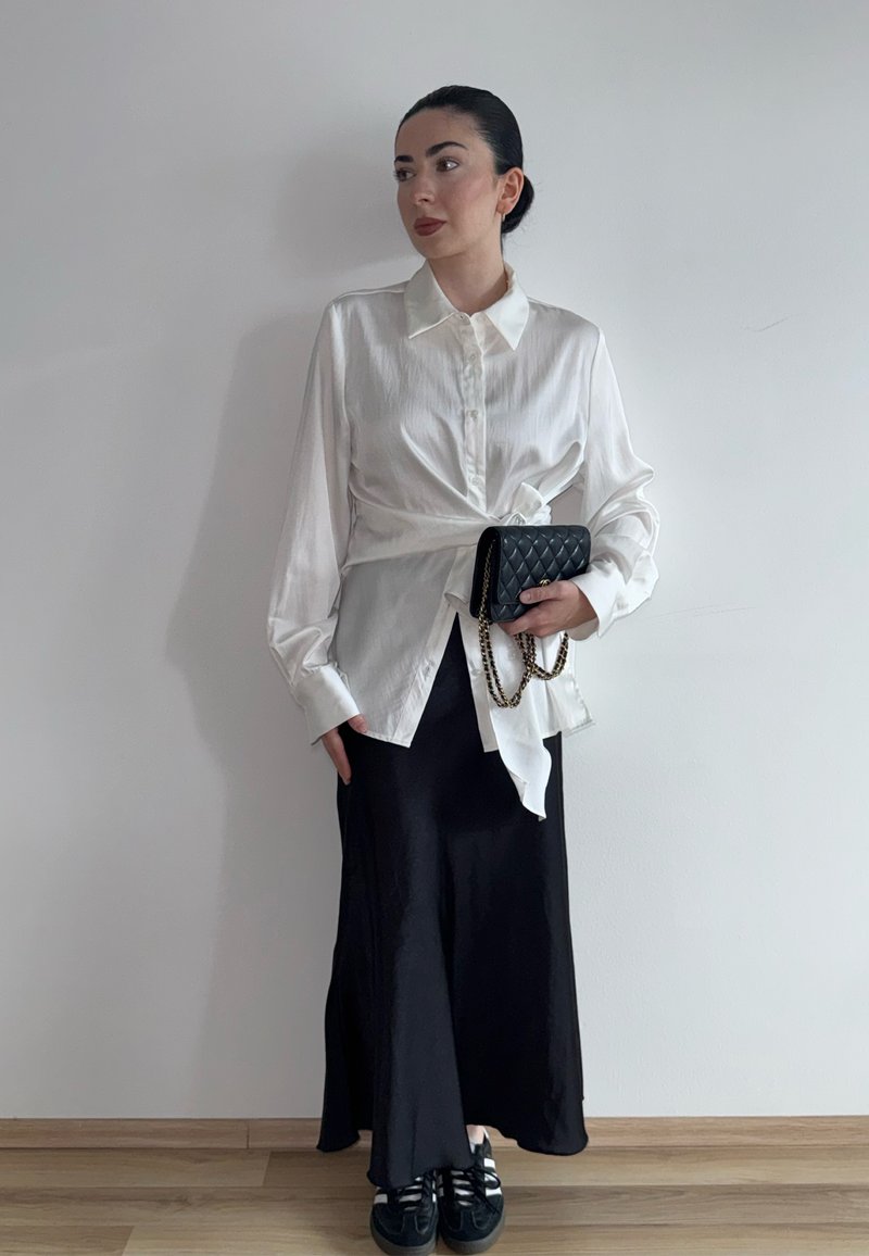 Femme aux cheveux foncés portant un chemisier blanc noué et une longue jupe noire, tenant un petit sac noir matelassé avec une chaîne dorée contre un mur blanc.