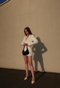 Hvit blazer over en nude topp, kombinert med svart håndveske og hvite spisse hæler. Langt, glatt hår og et naturlig sminkeutseende. Minimalistisk setting.