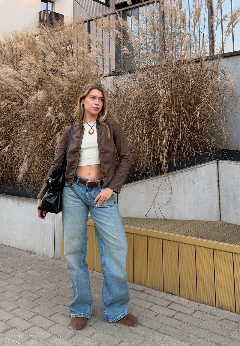 Jonge vrouw in een bruin jasje, een wit cropped topje en wijde jeans staat op een geplaveide stoep met hoge droge grassen achter haar.