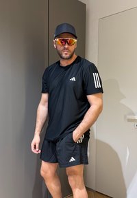 Homme portant une tenue de sport Adidas noire, une casquette noire et des lunettes de soleil orange réfléchissantes, debout à l'intérieur avec une main dans la poche.