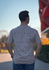 Light beige short-sleeved t-shirt featuring bold black text "CALVIN KLEIN JEANS" vertically aligned on the back, made of soft fabric.