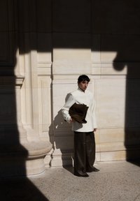 Kremfarget, tettsittende jakke, vide olivenbukser, holder en mørkebrun semsket skinnveske. Står mot en lys steinvegg med skygger.