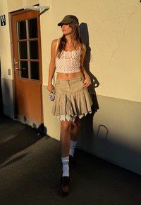 Top bianco senza maniche con pois rossi, gonna beige a pieghe, calze bianche e scarpe marroni. In piedi accanto a una porta, con la luce solare che proietta un'ombra.