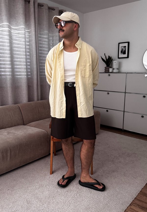 Man draagt een beige pet, zonnebril, geel open overhemd over een wit tanktopje, zwarte short en zwarte teenslippers, staand in de woonkamer.