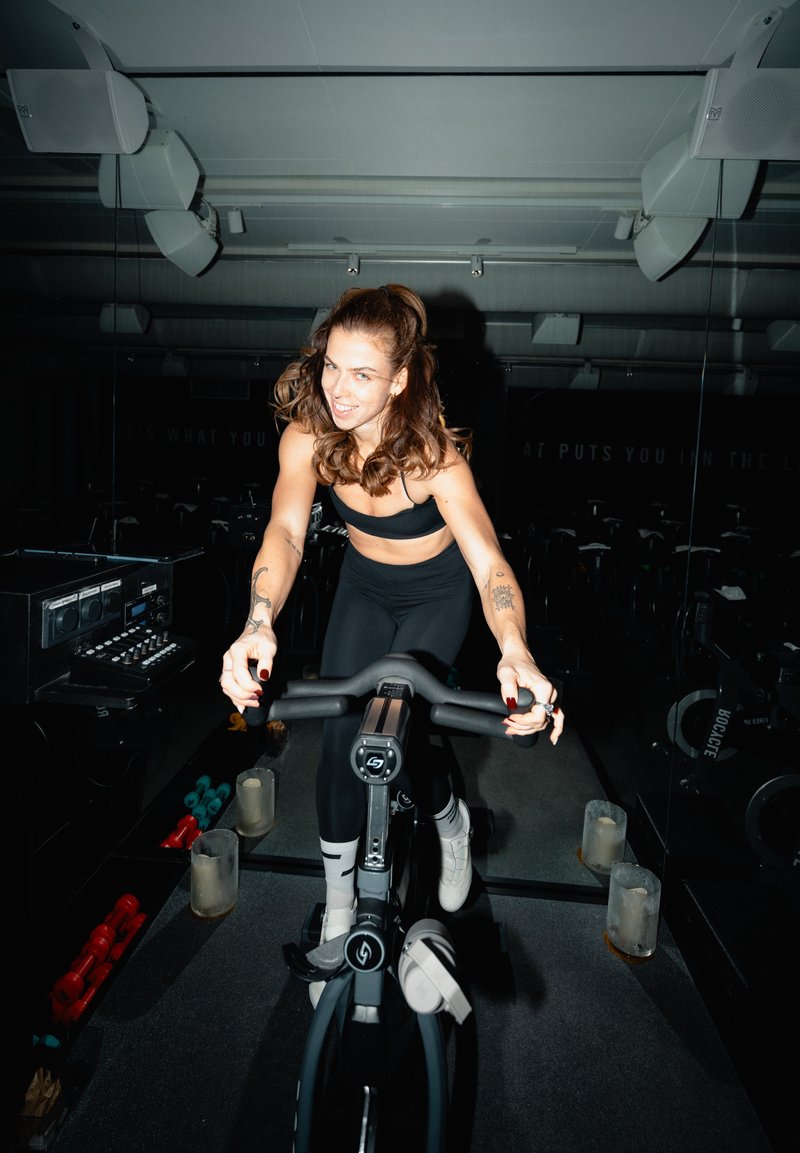 Kobieta z tatuażami w czarnym stroju sportowym jeździ na rowerze stacjonarnym w przyciemnionej siłowni, wokół znajdują się hantle i głośniki.