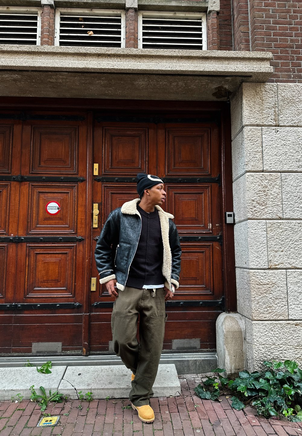 Fekete bőr bomberdzseki fehér gyapjú gallérral, sötét pulóver, olivazöld cargo nadrág és barna csizma, fa ajtók mellett állva.