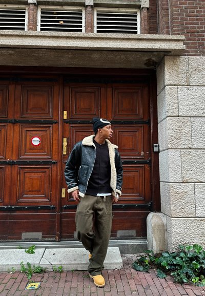 Chaqueta bomber de cuero negra con cuello de shearling blanco, suéter oscuro, pantalones cargo verde oliva y botas beige, de pie junto a puertas de madera.