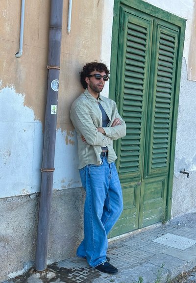 Camisa ligera a rayas, camiseta interior gris, jeans de pierna ancha azules y zapatos negros. De pie junto a una pared texturizada y contras de madera verdes.