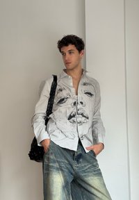 Camicia a maniche lunghe a righe con design grafico di un volto, abbinata a pantaloni in denim blu chiaro larghi. Borsa nera a spalla con tracolla borchiata.