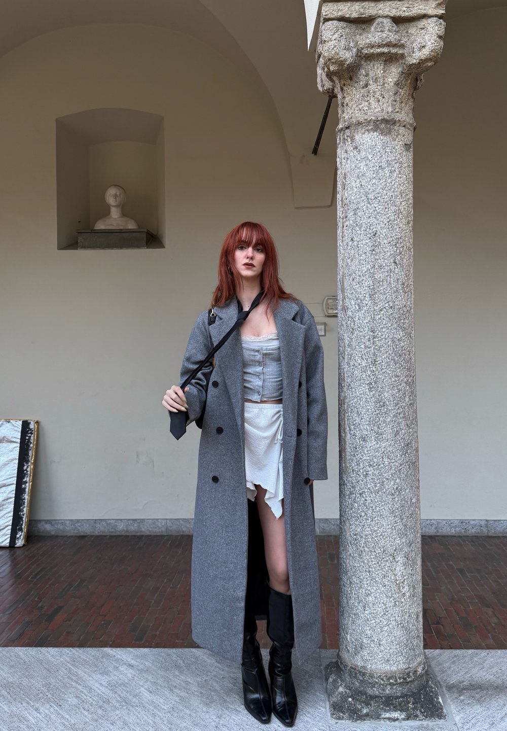Cappotto grigio in lana, top corsetto grigio chiaro, gonna bianca con volant e stivali neri fino al ginocchio. In piedi accanto a una colonna di pietra in uno spazio interno.