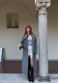 Cappotto grigio in lana, top corsetto grigio chiaro, gonna bianca con volant e stivali neri fino al ginocchio. In piedi accanto a una colonna di pietra in uno spazio interno.