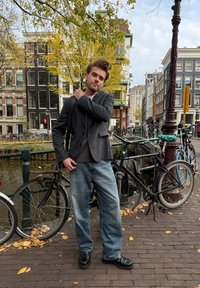 Homem vestindo um blazer escuro por cima de um suéter cinza, jeans azuis largos e sapatos pretos, está junto a bicicletas perto de um canal rodeado por folhas de outono.