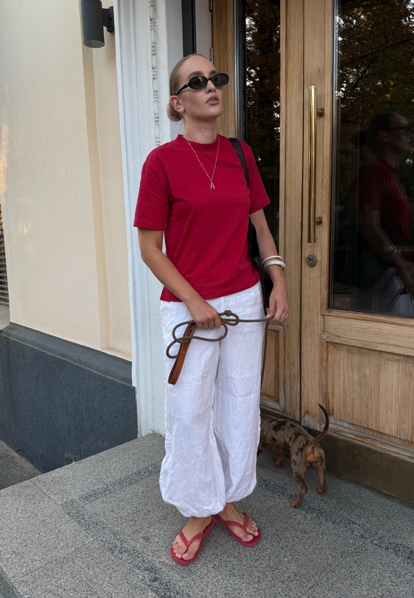 Femme en chemise rouge, pantalon blanc et sandales roses tenant une laisse près d’un petit chien brun près d’une porte en bois.