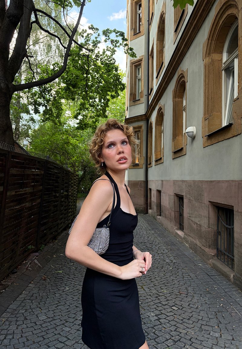 Femme aux cheveux bouclés portant une robe noire et un sac à motifs, debout sur un chemin pavé à côté d'un bâtiment avec des fenêtres en arc et des arbres au-dessus.