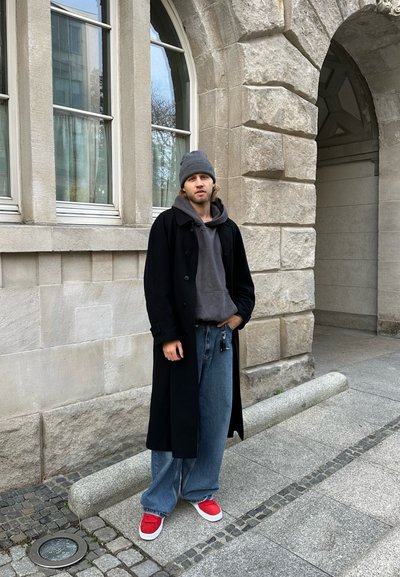 Abrigo largo negro, sudadera gris, vaqueros anchos azules y zapatillas rojas. Gorro gris. Pared de piedra y ventanas con arco de fondo.