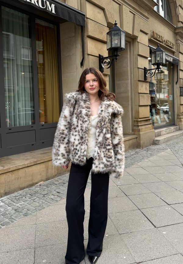 Ung kvinne som står på en fortauskant i byen, iført en flekkete fuskepelsjakke, hvit topp og svarte bukser, med vegglamper i vintage-stil i bakgrunnen.