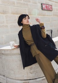 Femme en col roulé marron, manteau noir à fines rayures et ceinture léopard, appuyée sur un rebord en pierre, tenant une tasse de café près d'un panneau "METRO" sur un mur en pierre.