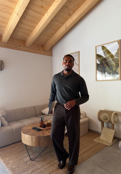 Hombre con suéter oscuro y pantalones sosteniendo una tableta, de pie en una sala de estar con techo de madera, mesa de centro redonda de madera y arte de plantas enmarcado.