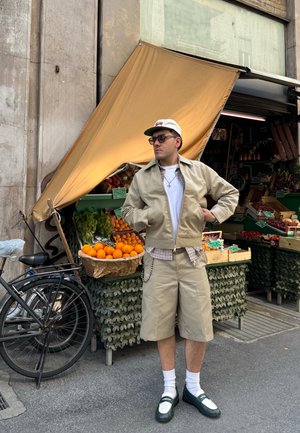 Chaqueta beige y pantalones cortos con bolsillos, camisa blanca, acento de cuadros, zapatos verdes, de pie frente a un puesto de frutas con naranjas y verduras.