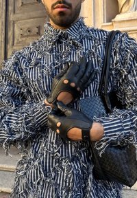 Homme portant une veste rayée noir et blanc texturée, des gants en cuir noirs sans doigts ajustant une main, tenant un sac à main noir tressé.