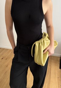Żółta skórzana torebka kopertowa z zebranym wierzchem, trzymana pod ramieniem. Założona z czarnym prążkowanym topem na ramiączkach i czarnymi spodniami w prążki.