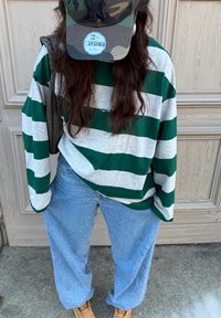 Chemise à manches longues rayée verte et grise associée à un jean large texturé bleu clair et des bottes beige. Une casquette camouflage complète le look.