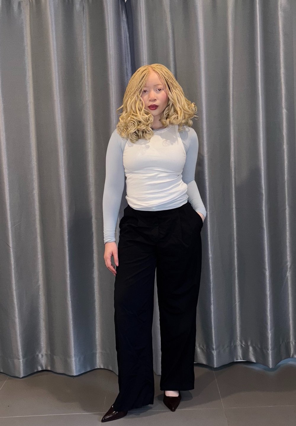 Haut gris clair à manches longues associé à un pantalon noir ample, posé devant un rideau gris. Tissu lisse avec une texture subtile.