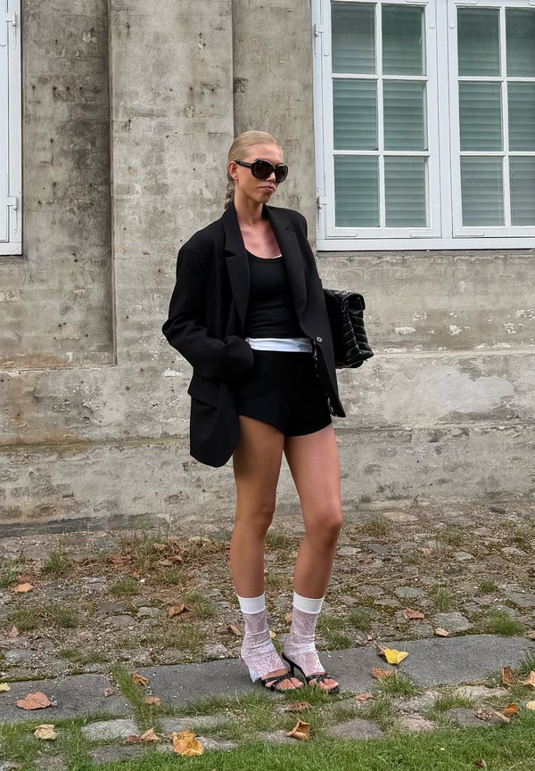 Ženska z blond lasmi v čopku, oblečena v prevelik črn blazer, črne kratke hlače, črn top, bele čipkaste nogavice, črne pete in sončna očala, stoji zunaj.