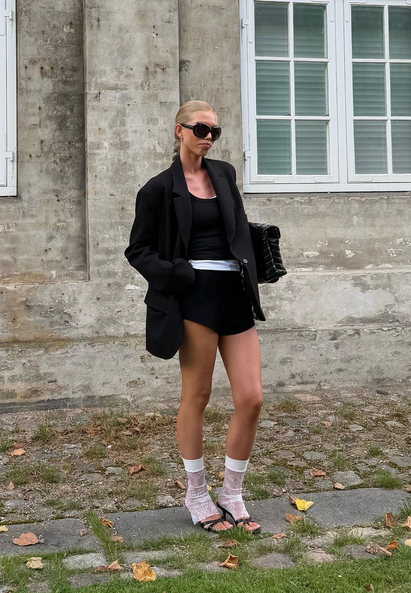 Ženska z blond lasmi v čopku, oblečena v prevelik črn blazer, črne kratke hlače, črn top, bele čipkaste nogavice, črne pete in sončna očala, stoji zunaj.