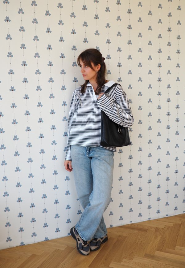 Mujer con camisa de manga larga a rayas y vaqueros, de pie frente a una pared con estampado floral, con bolso negro de hombro y deportivas sobre suelo de madera.