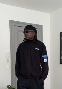 Homme portant une veste Adidas noire, une casquette noire et des lunettes de soleil, debout à l'intérieur avec les mains dans les poches près d'une porte grise et d'un mur blanc.