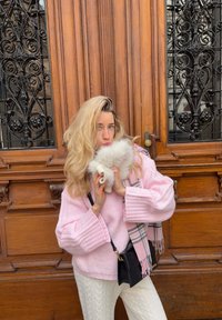 Pull rose en tricot avec manches larges, pantalon blanc texturé et écharpe rayée ; tenant un accessoire blanc duveteux devant un fond en bois.