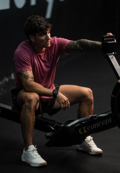 Máquina de remo Concept2 con un marco negro. Una persona con una camiseta rosa y pantalones cortos está sentada en ella, vistiendo zapatillas deportivas blancas y un reloj.