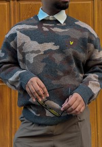 Homme portant un pull à motif camouflage sur une chemise blanche à col, tenant des lunettes de soleil noires avec des verres jaunes devant une porte en bois.