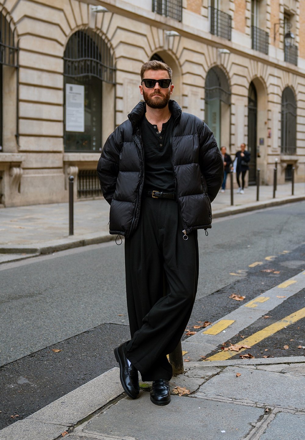 Doudoune noire à col montant, chemise noire à manches longues et pantalon large noir. Lunettes de soleil noires et chaussures noires polies.