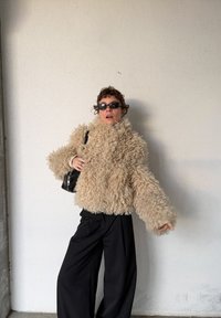 Personne portant une veste en fausse fourrure beige bouclée, un pantalon noir à jambes larges, des lunettes de soleil noires, et tenant un petit sac noir, devant un mur uni.