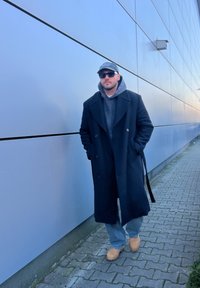 Μαύρο διπλό μπλέιζερ πάνω από μια γκρι φούτερ, σε συνδυασμό με ανοιχτό μπλε τζιν και καφέ ξύλινες παντόφλες, που στέκεται μπροστά από έναν ανοιχτό μπλε τοίχο.