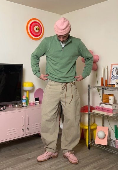 Suéter verde con cremallera, pantalones beige holgados con cordón y mocasines rosas. Los accesorios incluyen un gorro rosa, con decoración colorida de fondo.