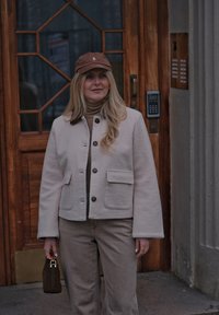 Femme portant une veste et un pantalon beiges, une casquette marron et un petit sac à main, debout devant une porte en bois avec un panneau d'interphone.