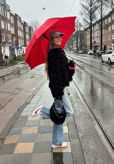 Paraguas rojo, chaqueta negra de lana, gorra gris, guantes negros, vaqueros azules, zapatillas blancas con acentos rojos, de pie en una acera mojada en un entorno urbano.