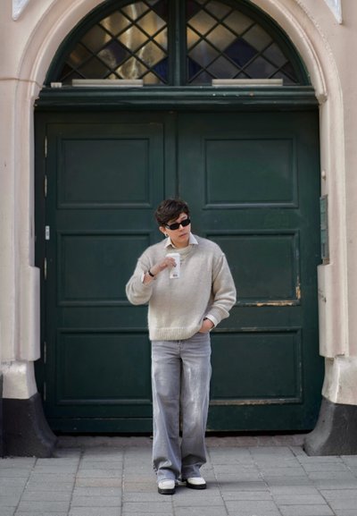 Suéter de punto gris claro con puños enrollados, jeans gris claro y mocasines negros. Sosteniendo una taza blanca, de pie frente a una puerta verde.